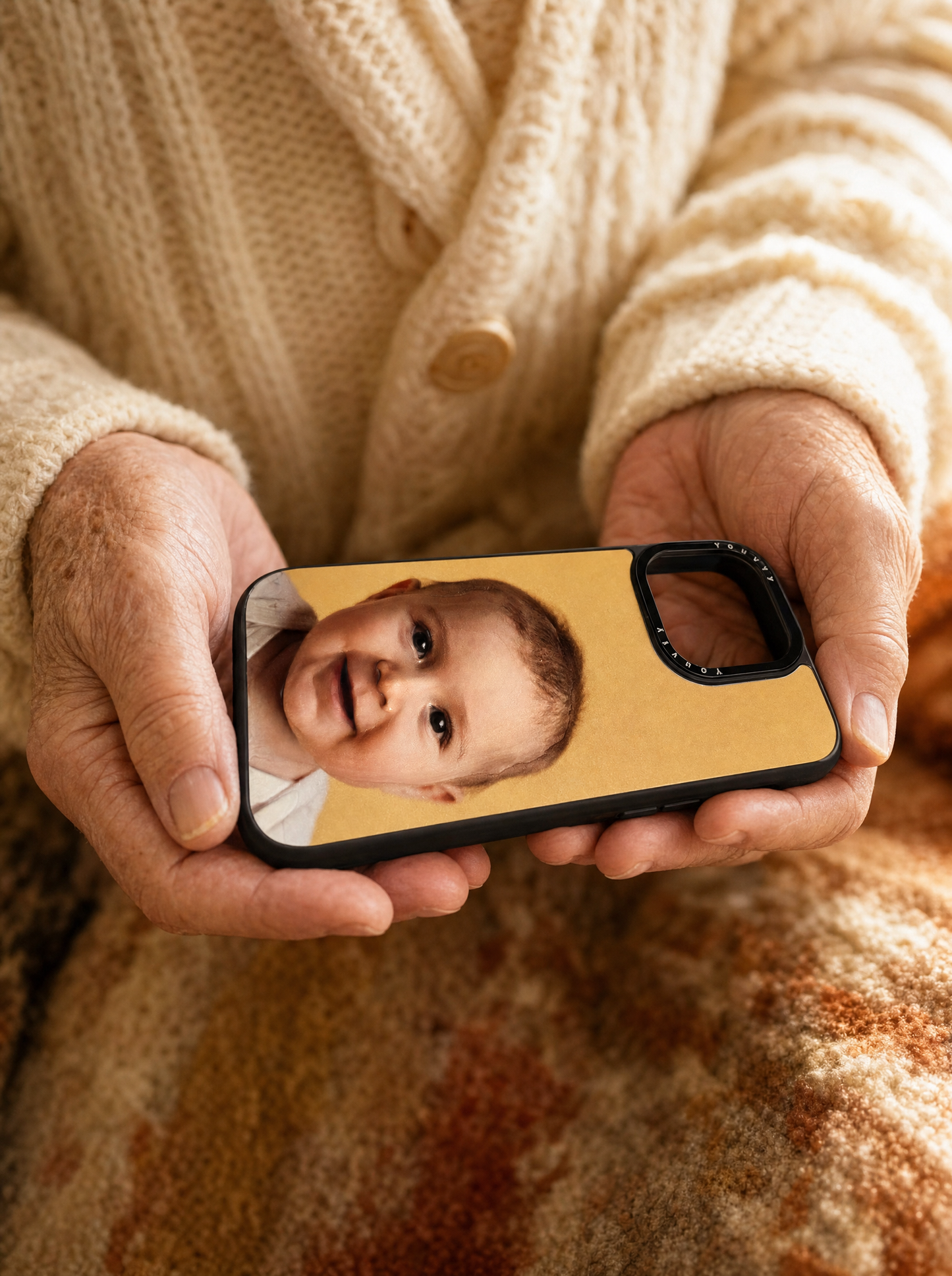 Older woman's hands cradling a Youvfy iPhone case with a baby grandchild portrait, warm cozy palette