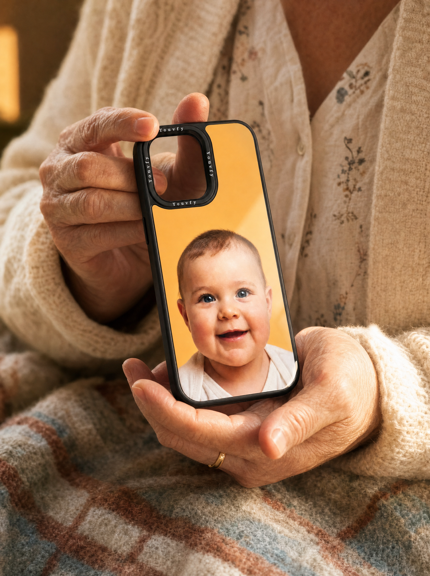 Older woman's hands cradling a Youvfy flat-printed iPhone case with a baby grandchild portrait, warm cozy palette